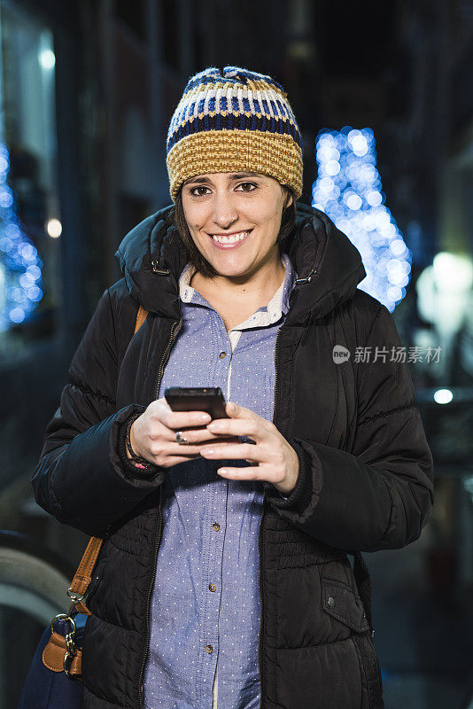 漂亮的女人在圣诞夜用智能手机看着摄像头，背景是装饰灯
