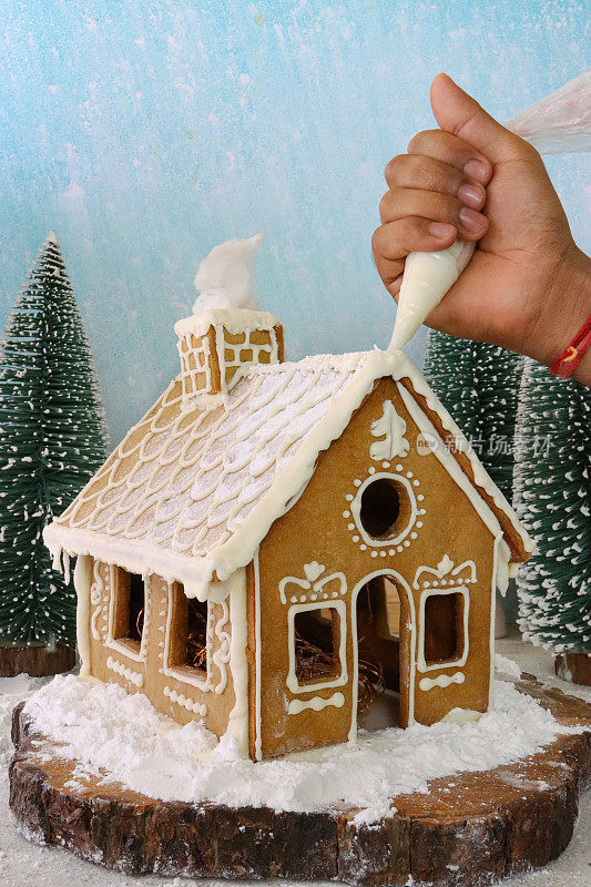 自制的姜饼屋被不认识的人用滚边袋的白色皇家糖霜装饰，展示在白雪覆盖的针叶林场景中，糖霜糖雪，模型冷杉树
