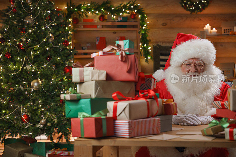 圣诞老人坐在办公桌前，桌上放着一堆礼物，我们看到他坐在装饰好的办公室里的礼盒后面，背景是圣诞树
