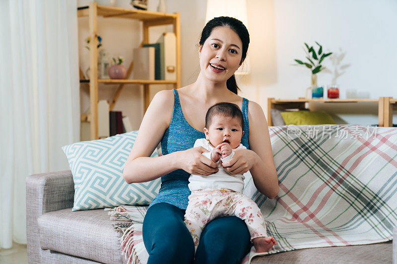 微笑的日本女士坐在沙发上和她的宝贝女儿。美丽的亚洲妈妈面对镜头，握着孩子的手。