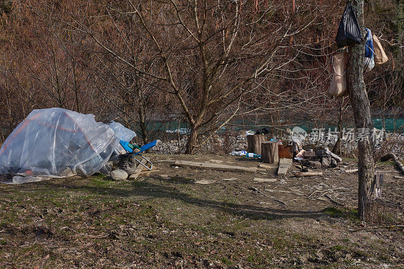 把无家可归的人停在山间河流岸边的森林里。帐篷被保鲜膜覆盖着。靴子踩在泥里。一袋袋的食物挂在树上。