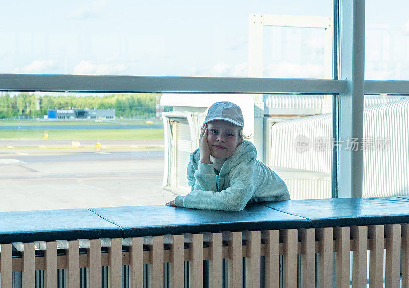 一个孩子，一个白人女孩在机场一个巨大的窗户的背景下看着相机镜头。