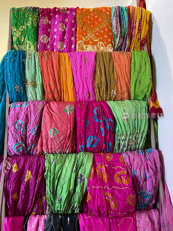 近距离的图像，一排排充满活力的，五颜六色的折叠织物，市场摊位展示，民族，传统服装挂在木架上，头巾和纱丽，重点在前景