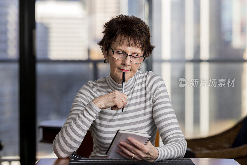 一位年长的女士坐在家里的桌子旁，拿着笔和记事本，一副若有所思的表情