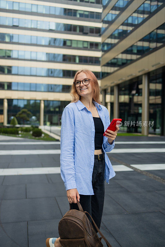 快乐的年轻金发女子在漫游中打电话的垂直镜头穿着休闲的衣服有高兴的表情走在模糊的城市建筑享受智能手机通话