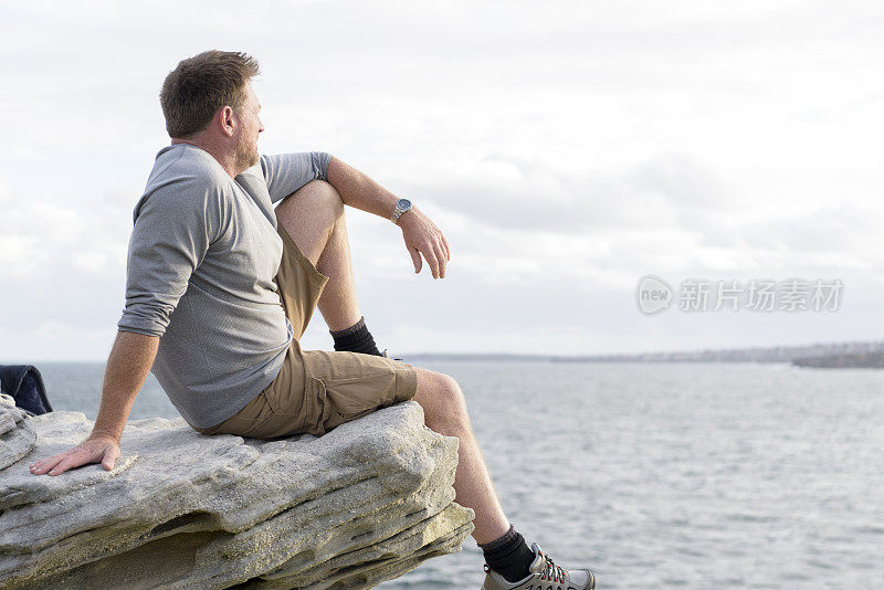 一个成熟的男人坐在岩石上看海，邦迪海滩