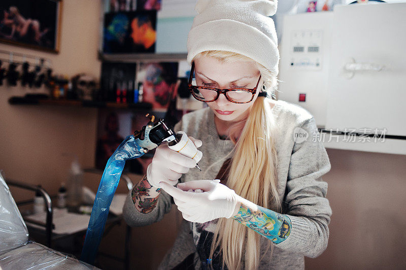 一个年轻的金发女孩的肖像大师纹身在眼镜和帽子与纹身机的手