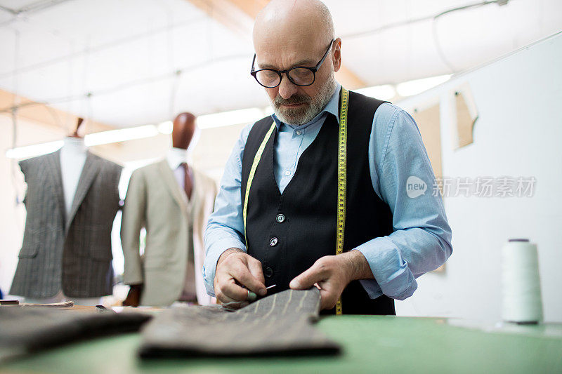 严肃忙碌的秃顶胡子男裁缝戴着眼镜，穿着时髦的衬衫和马甲，用针缝纫织物，在桌子上同时生产男装