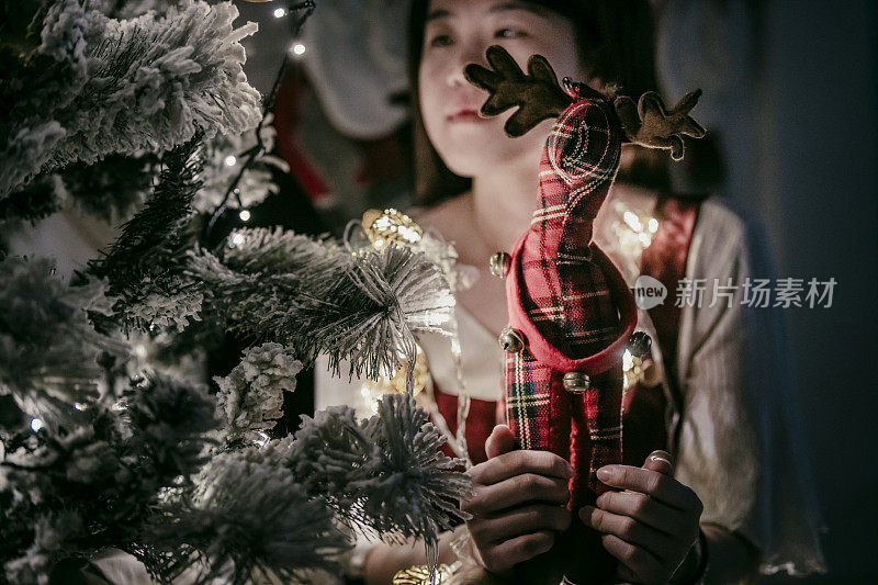 一名亚裔华人女性拿着毛绒玩具坐在圣诞树前，看着它