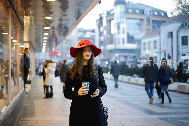 年轻女子戴着红帽子在街上一边喝着咖啡一边用智能手机发短信