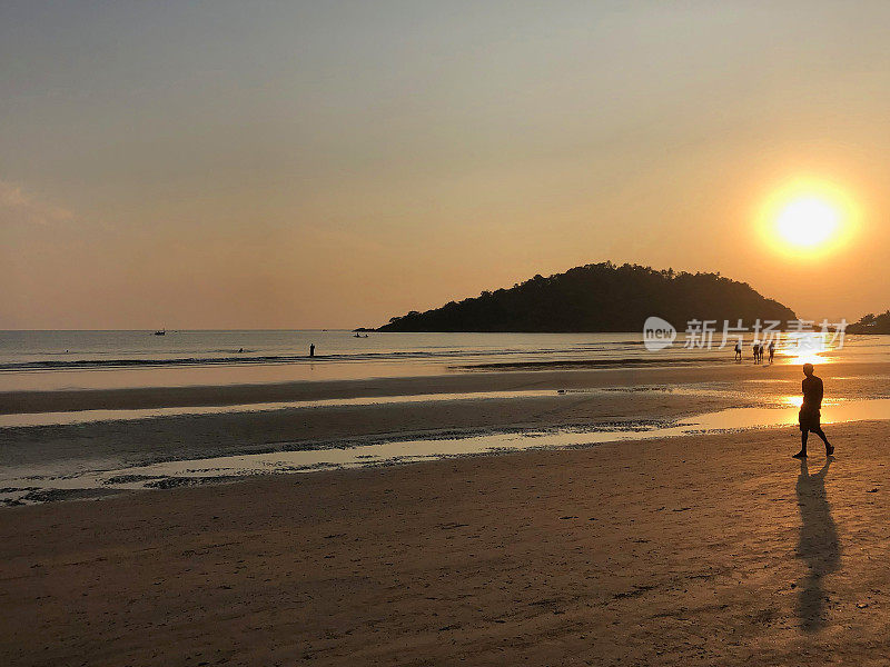 图片中，一名穿着短裤的男子走在印度果阿的帕洛伦海滩的海岸线上，夕阳下，海浪拍打着海滩