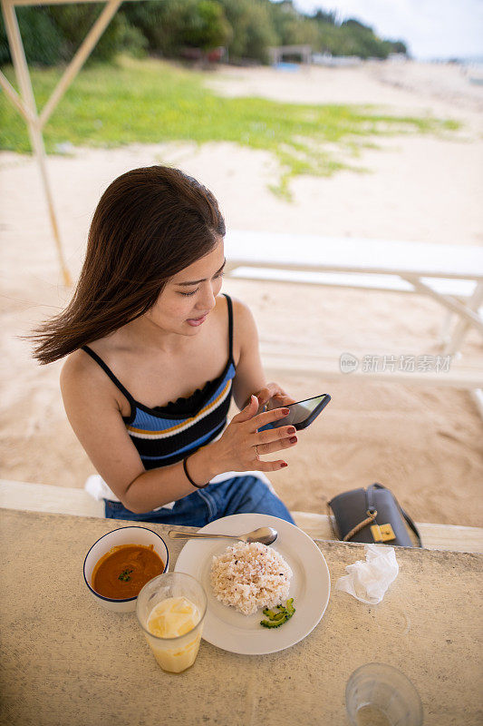 年轻女子使用智能手机在度假海滩午餐