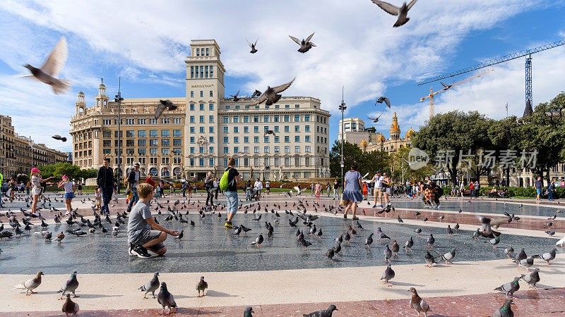加泰罗尼亚广场——在一个阳光明媚的秋夜，游客们在西班牙加泰罗尼亚的巴塞罗那市中心的加泰罗尼亚广场上与一大群鸽子嬉戏。