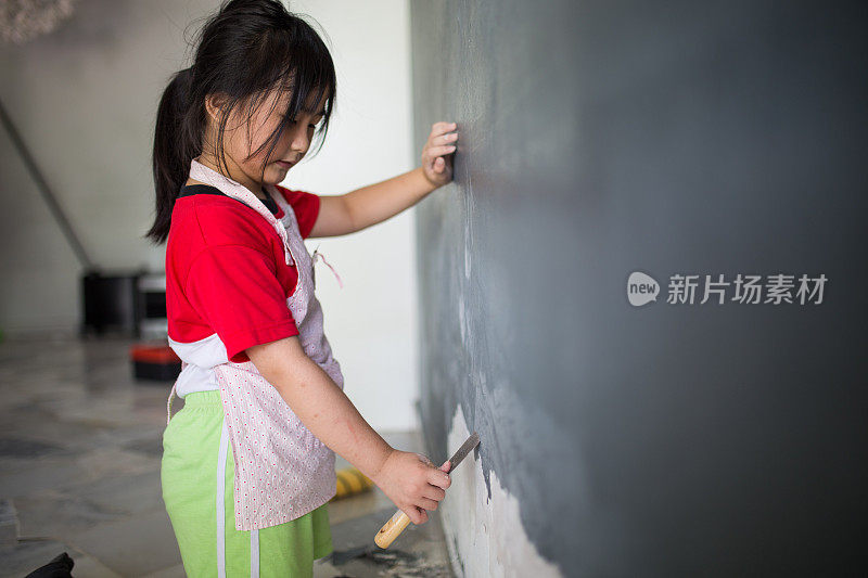 一个亚洲中国女孩正在帮助家人刮掉墙上脱落的部分，为粉刷做准备