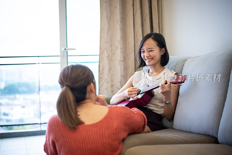 千禧一代的年轻亚洲女性与她最好的朋友在她的公寓里弹奏她的尤克里里琴。