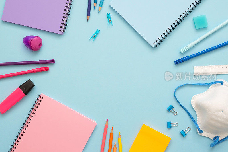 以蓝色为背景的彩色文具。俯视图蓝色，粉色和紫色的笔记本，回形针，橡皮，毡尖笔，彩色铅笔，马克笔和口罩。