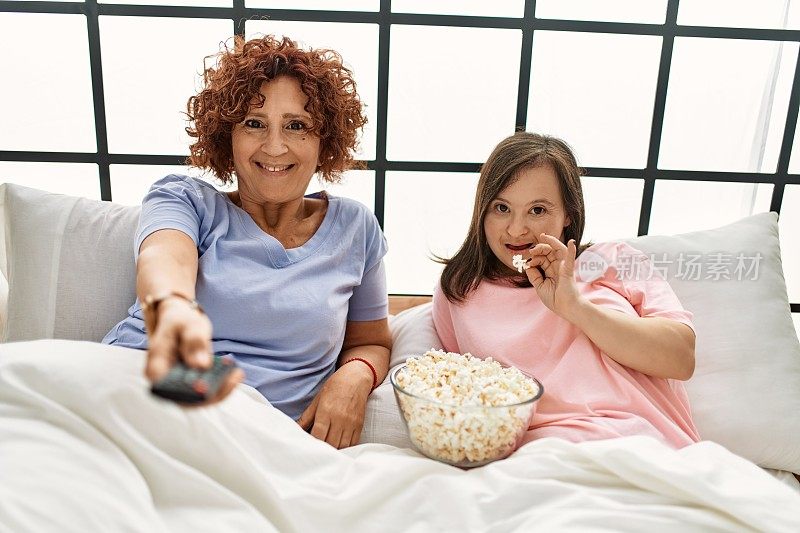 母亲和唐氏综合症的女儿舒舒服服地躺在床上，吃着爆米花，看电视
