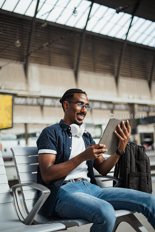 一个快乐的非洲人坐在火车站的长凳上使用平板电脑