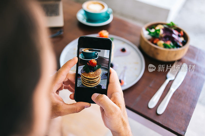 男性影响者通过智能手机摄像头为社交网络拍摄食物的裁剪图像。博主拿着手机拍下咖啡馆早午餐上摊着的美味煎饼