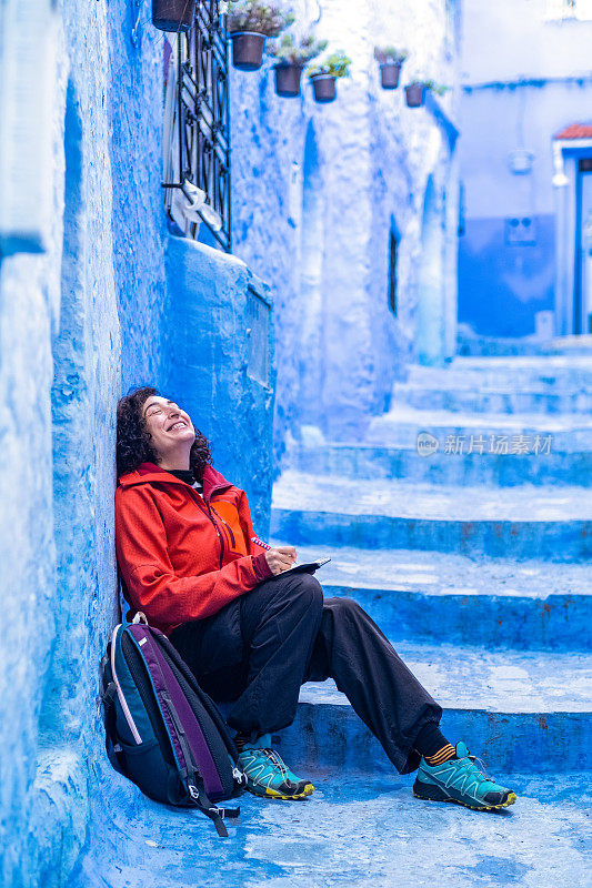 一名独自旅行的女旅行者，穿着红色夹克，摆出幸福和幸福的姿态，坐在蓝色房屋的旅游小镇的楼梯上，在记事本上写字。