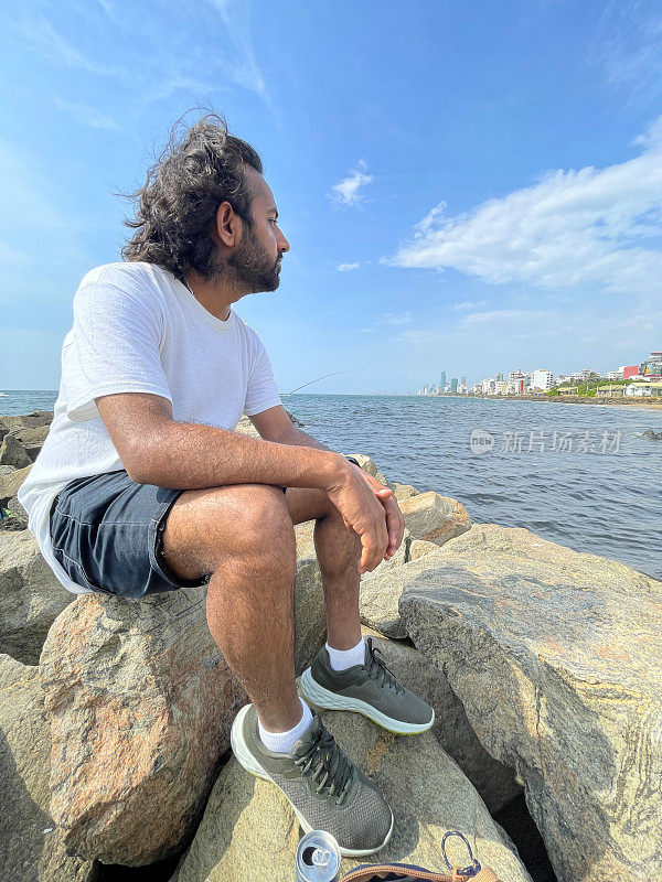 特写图片，印度人坐在斯里兰卡海岸线上的人造岩石海防上，看着风景，海岸侵蚀抛石装甲墙，海岸管理，大海，背景是斯里兰卡科伦坡的城市摩天大楼
