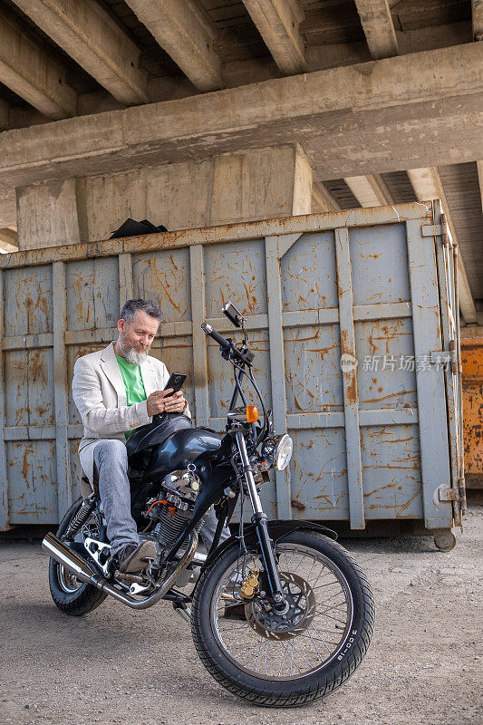 一个成熟的男人，穿着浅绿色的t恤，米色的夹克和灰色的牛仔裤，骑在一辆摩托车上。
