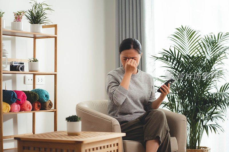 一名亚洲女性坐在客厅的扶手椅上使用智能手机，患上了眼睛疼痛。