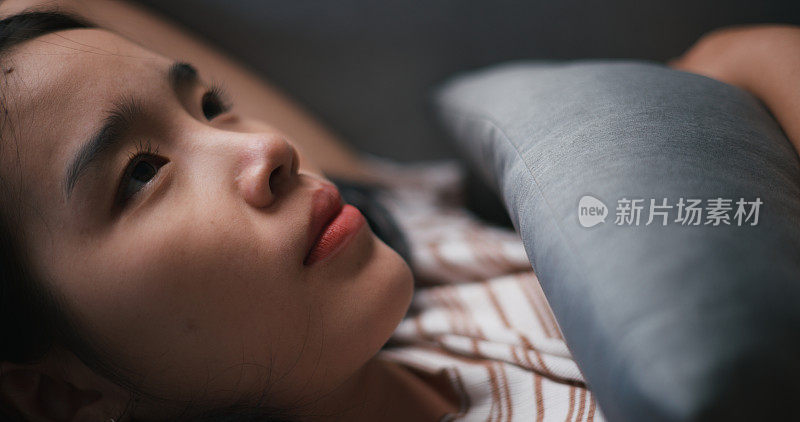 忧郁沮丧的年轻亚洲女子躺在家里的沙发上，看着天花板思考着