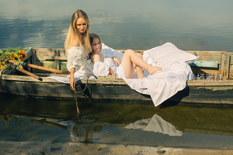 两个穿白衣服的金发女人在湖边度假。女性的友谊。旧渔船上的白色亚麻布和桨。床单。夏天放松