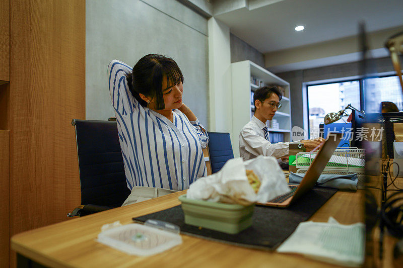 一位年轻的亚洲白领女性住在繁忙的办公室里。在午休时间，年轻的办公室女性正在电脑前做一些简单的伸展运动，以缓解长时间坐在电脑前的不适