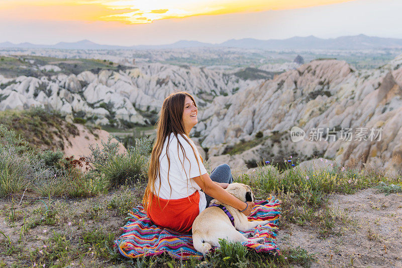 快乐的女人和她的狗在土耳其卡帕多西亚风景秀丽的山谷里欣赏日落