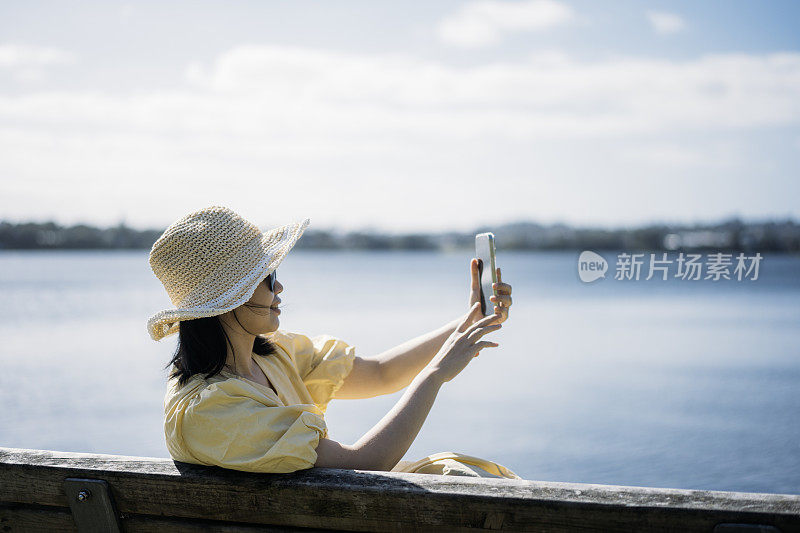 年轻的亚洲女子坐在湖边的长凳上，用智能手机自拍