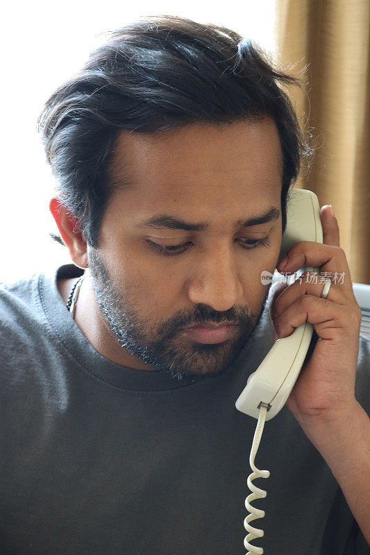 全画幅的印度男子穿着休闲服在办公室工作，听着座机电话，把电话听筒举到耳边，头像，焦点在前景