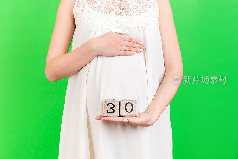 近距离的肖像孕妇穿着白色礼服显示立方体怀孕30周在绿色背景的腹部。宝贝期待。本空间