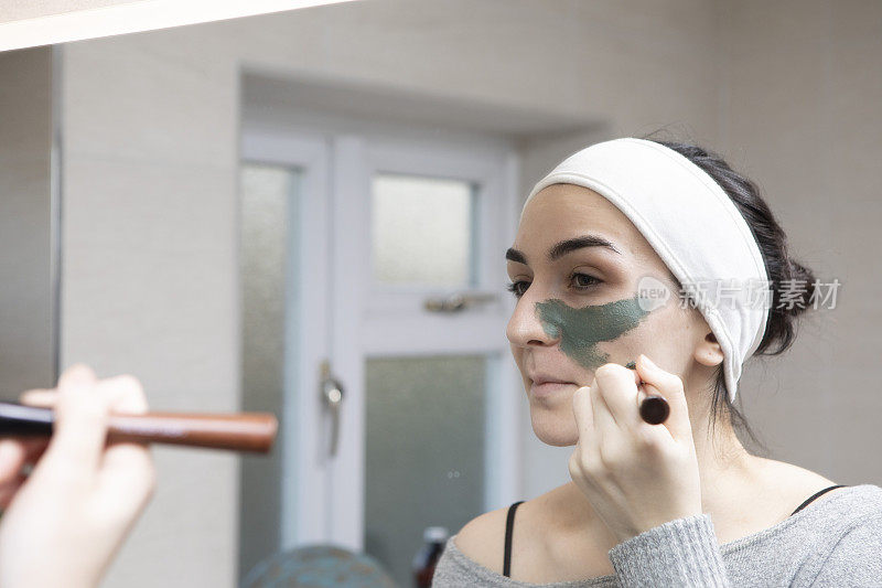一名年轻女子在家用浴室的镜子前用刷子涂绿色面膜的肖像