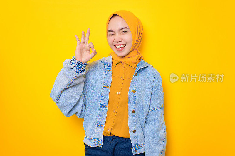 微笑的年轻亚洲妇女在牛仔裤夹克显示okay手势，表明认可孤立的黄色背景