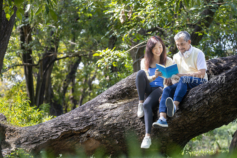 亚洲高年级的父亲和女儿在夏天一起坐在成熟的树枝上阅读一本书，以身心健康和长寿的概念