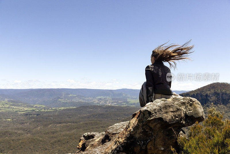 年轻女子坐在岩石上，在刮风的日子，看着山上山谷，背景与复制空间