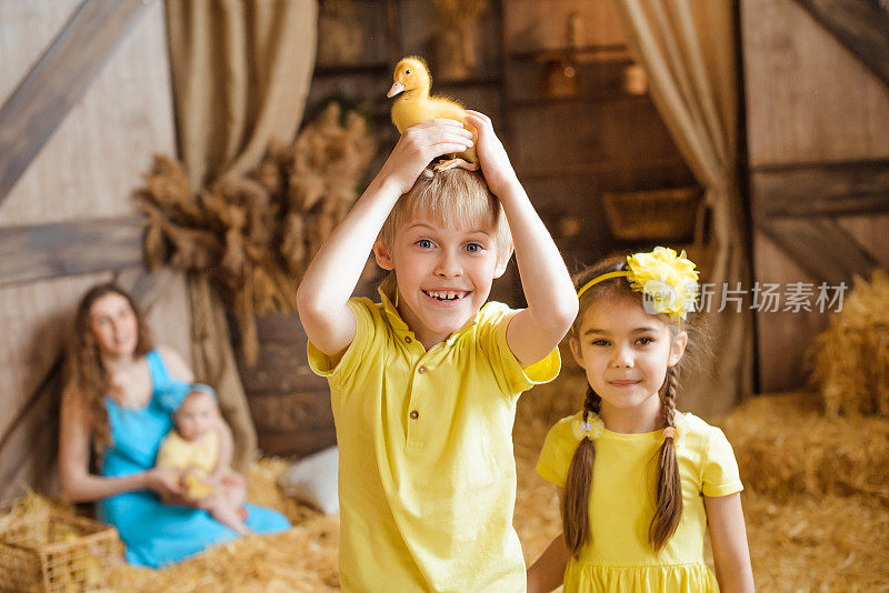 男孩的头上顶着一只黄色的小鸭子，他的妹妹站在他旁边，两人都笑了。他们站在一个放着干草捆的农村谷仓前。
