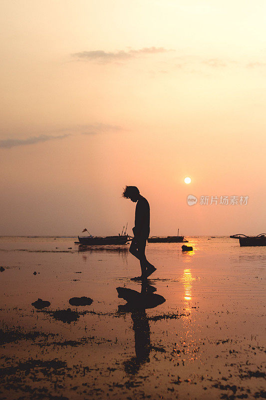 一个人在日落时走在海滩上的船旁边