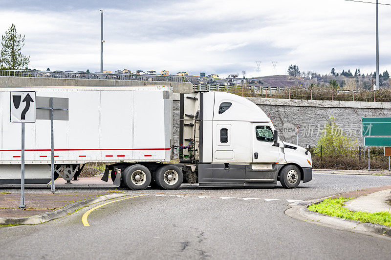 一辆白色大钻机半挂车的侧视图，用干式货车半挂车运输货物，行驶在圆形交叉路口的当地道路上