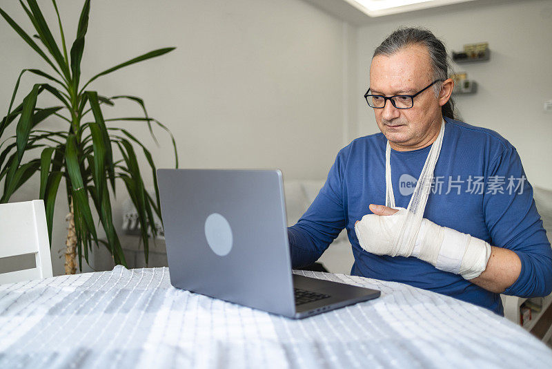 中年白人男子手臂骨折，打了整形石膏，在家用笔记本电脑