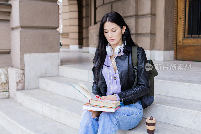 年轻漂亮的女大学生背着双肩包坐在大学楼梯上。她戴着耳机，穿着皮夹克，等待着大学课程的开始。