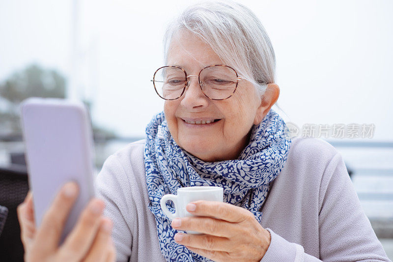 一位面带微笑的白发老妇人坐在户外的咖啡桌旁，看着智能手机上的社交应用，喝着浓咖啡休息一下