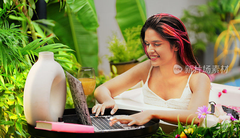 印度美女坐在浴缸里，用笔记本电脑打字，看着屋外的屏幕。快乐的年轻成人z世代微笑的女孩在电脑上聊天网上约会享受豪华酒店度假别墅之旅