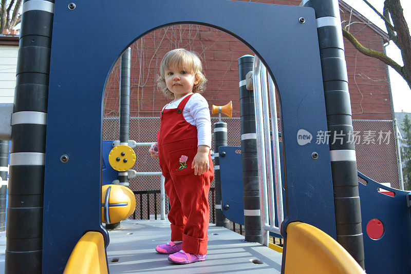金发的蹒跚学步的女孩穿着红色的工装裤在滑梯上。