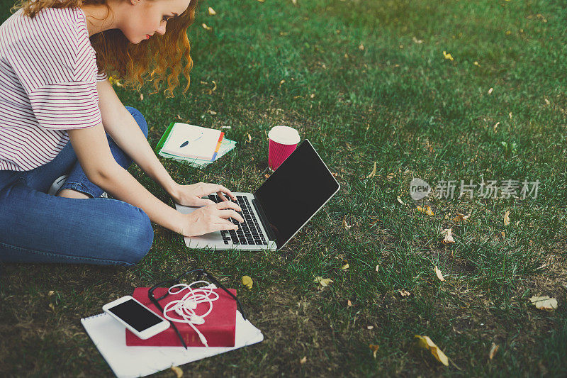 年轻的红发女孩坐在树下随意使用笔记本电脑