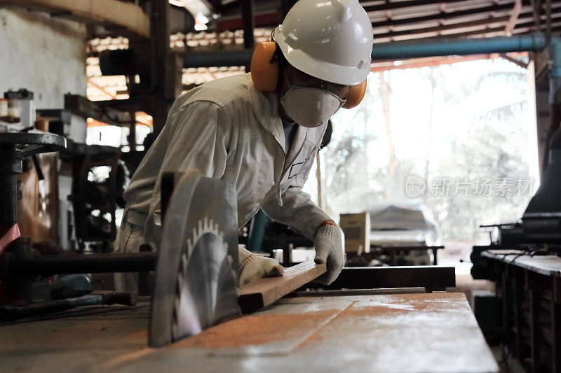 在木匠厂，穿着白色制服和安全设备的专业年轻工人在台式锯机上切割一块木头。