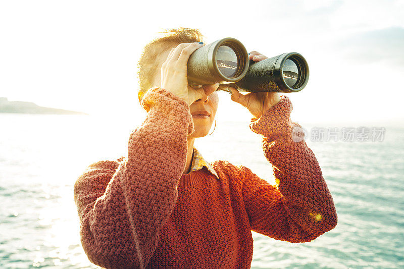 在一个阳光明媚的日子里，美丽的年轻女孩通过双筒望远镜看大海。旅游热旅程的概念
