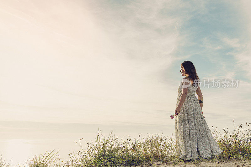 一个穿着夏装的女人站在沙丘上看日落，康沃尔